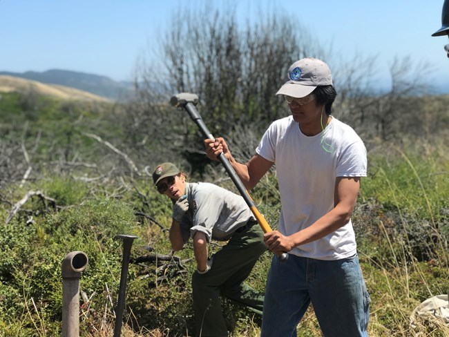 A young man swings a sledgehammer at a vertical pipe as a National Park employee in the background looks on.