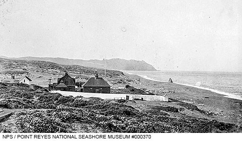A black and white photo of several wooden structures adjacent to an ocean beach on the right with rugged headlands rising in the distance.