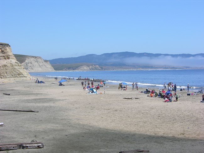 Dozens of visitors on a sandy beach with bluffs rising on the left and in the distance.