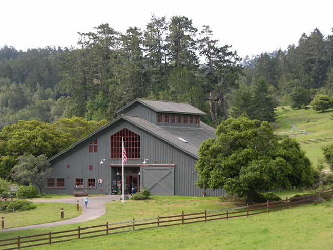Visitors entering and exiting a gray barn-like visitor center surrounded by green pasture and trees.