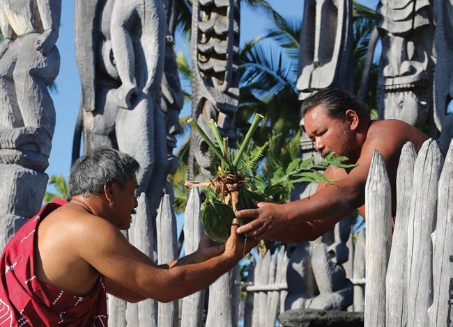 A man in traditional clothing hands a hoʻokupu (offering) to another man in traditional clothing at Hale o Keawe