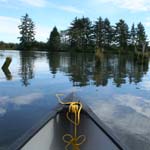 Bow of a canoe floating over the water reflecting the shore and sky