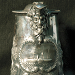 A detail of the front of a silver pitcher with a detailed relief image of a steamship.