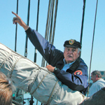 A volunteer docent leaning on a furled sail aboard BALCLUTHA and speaking to visitors.