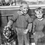A black and white photo of two boys and a dog standing on the deck of a 19th century sailing ship.