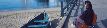 A visitor on Hyde Street Pier sits on a bench near a rowboat on the beach near the water.