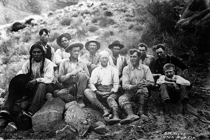 Historical photo of a group of men sitting on the ground in scrubby desert brush and rocks.