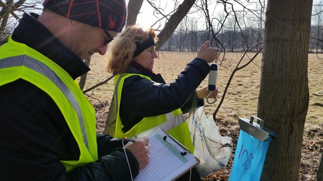A woman prepares to weigh a bag of maple sap while a man notes the tree number on a data sheet.