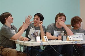 Four students in matching gray shirts sit at a table; two "high-five" each other.
