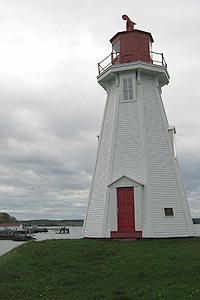 White and red lighthouse sits on a grassy area above a bay.