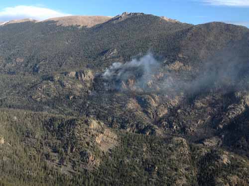 Black, burned area with smoke rising from middle. Rocky, mountainous terrain. 