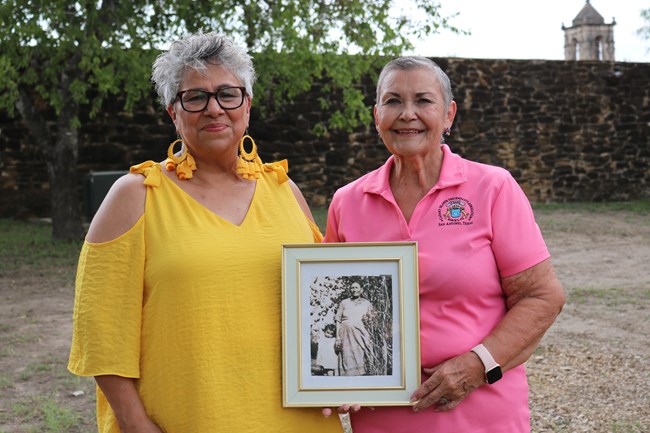 Two mission descendent women standing in front of Mission San Jos&#é;ssion descendent women standing in front of Mission San José holding a framed picture of their ancestor.