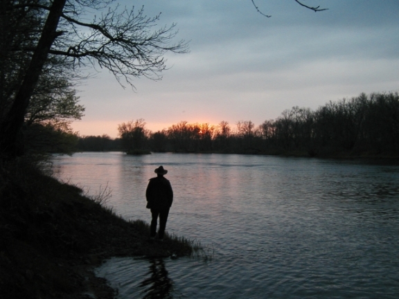 A person stands next to the St. Croix at sunset in spring silhouetted against the moving water and treeline across the river. (NPS Photo)