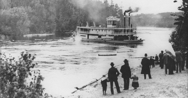 Black and white photo of a steamboat in a river. People standing on shore looking at it.