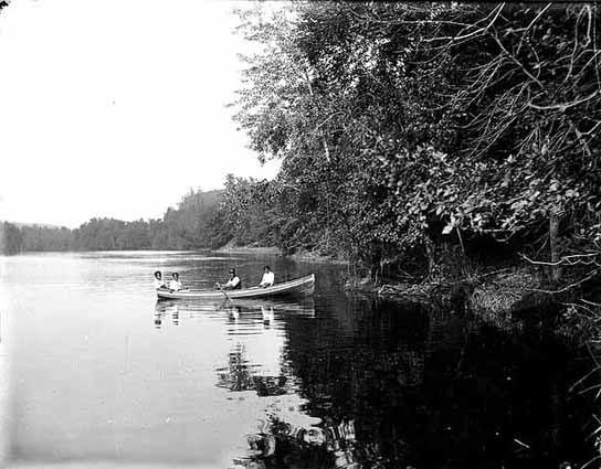 A black and white image of four people in a row boat along tree lined river