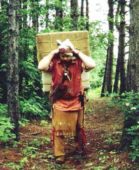 Man dressed in leggings and shirt with sash, walks a trail carrying a large square pack on his back with strap around forehead