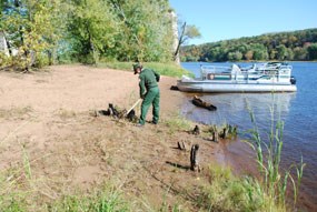 A uniformed park ranger rakes the sand.  A pontoon boat is tied up to the shore. A tree line is in the background.
