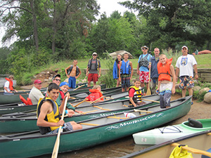 A group of youth and adults with canoeing equipment stand next to a river.