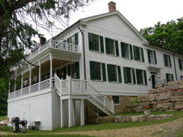A two story white house with green shutters.  Facing the river is a two story porch.
