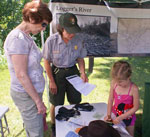 A ranger and a female adult look on as a child stamps a piece of paper.  A historic photo and map are in the background 
