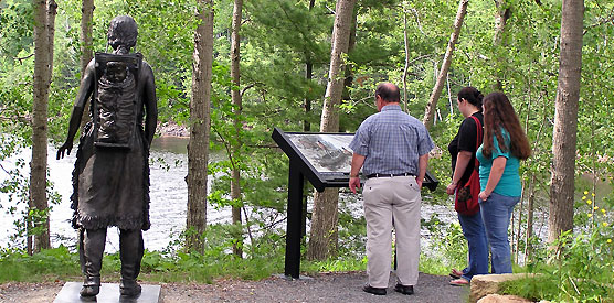 Two women and a man look at a wayside exhibit next to a bronze statue