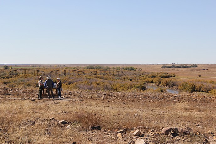three people standing around a wayside exhibit situated in an open prairie along a dirt trail, frame of small metal bridge in distance