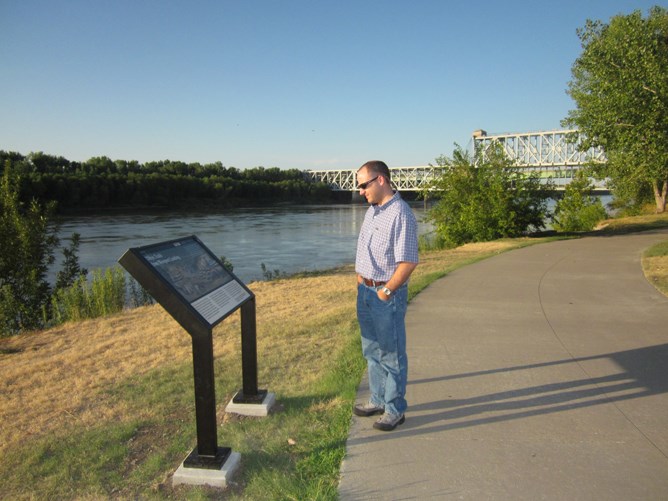 man on paved path reading wayside exhibit along river, metal bridge in the background