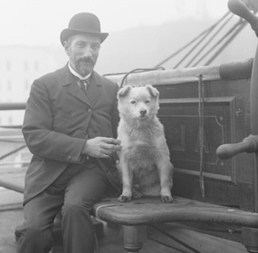 A man in a bowler hat seated next to a white dog near the helm of a sailing ship. A man in a bowler hat seated next to a white dog near the helm of a sailing ship.