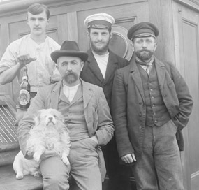 Four men and a white dog posing on the deck of a 19th century sailing vessel.