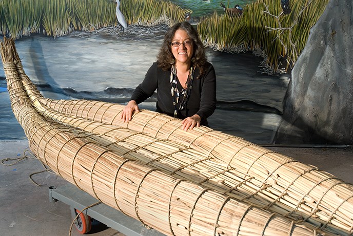 A woman sitting behind a canoe made out of tule reeds. She has her hands resting on the tan-colored canoe.