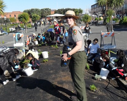 A park ranger standing in the middle of an empty garden plot with kids planting native plants behind her.