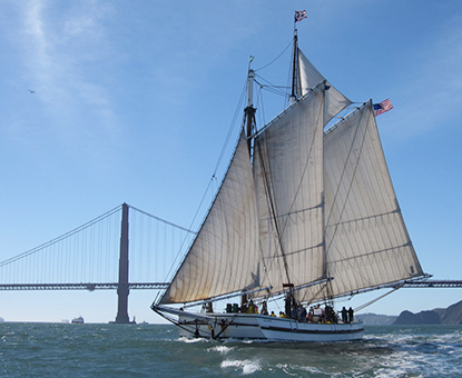 Scow schooner ALMA with her sails raised on San Francisco Bay with the Golden Gate Bridge in the background.