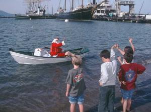 Children standing on a beach waving to Santa Claus in a rowboat with Hyde Street Pier in the background.