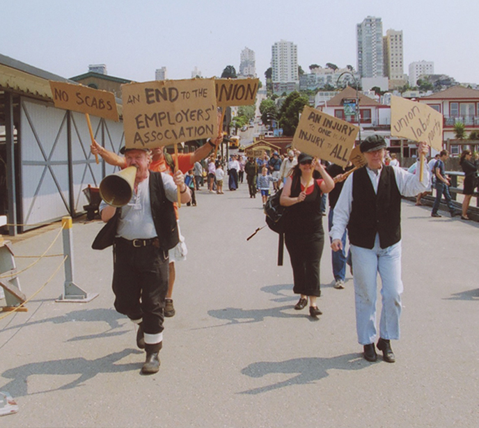 Living History players dressed in 1901-era clothing waking on the pier carrying signs.