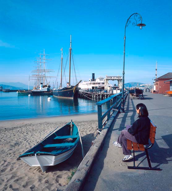 A woman sitting on a bench near a small beach with Hyde Street Pier in the background.