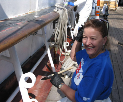 A smiling woman in a blue tee shirt holding a paint brush on the main deck of the BALCLUTHA.