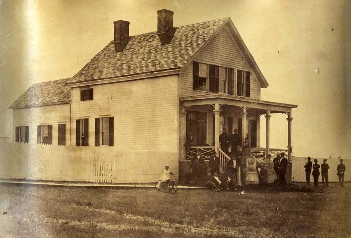 black and white photograph of a frame house with two chimneys and a large group of people gathered on the porch.