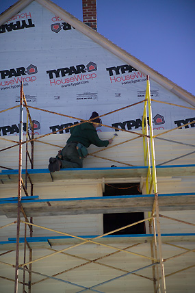 workman on a scaffold nailing clapboards to one of the store house's gables.