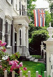 The front of the Stephen Phillips House, a gray three story building with black shutters and a white portico.  The Hawaiian flag hangs in front of the house.