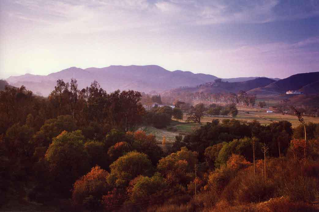 The Gillette Ranch valley seen at dusk in the autumn.  Orange, brown and yellow trees highlight the valley with white Spanish Revival buildings peaking above the trees in the valley below the mountains.