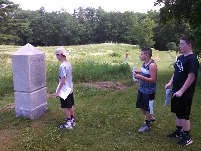 Three students stand by a monument in the park filling out worksheets.