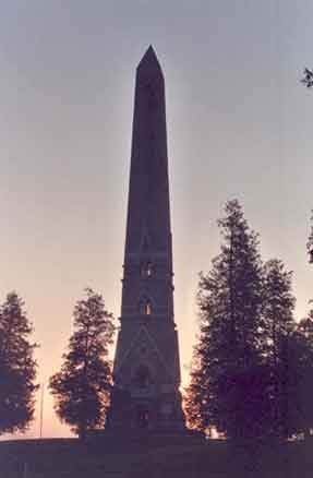 View of Saratoga Monument at sunrise.  The dark shadow of the Monument rises into the early morning sky, the scene lightly backlit by the not yet risen sun.