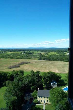 View from the top of Saratoga Monument, with clear, deep blue skies, rich green trees and lawn, light brown farm fields, and faded green hills in the distance.