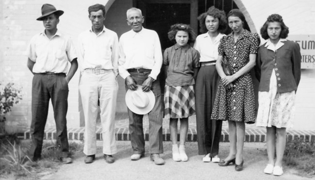 A family of seven people stands in front of the entrance to a building.