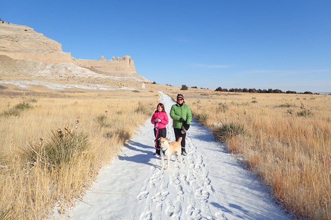 A woman and young girl walk a snow-covered trail with their dog on a leash