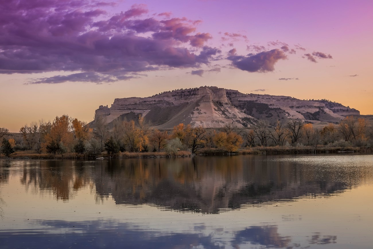 A purple and pink sky is seen above a sandstone bluff and is reflected in the waters of a river.