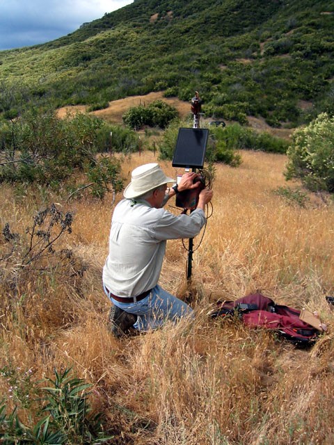 Scientists kneels in front of recording equipment and a small solar panel mounted to a pole to record bat calls in an area of grasslands and shrubs in the Sequoia National Park foothills.