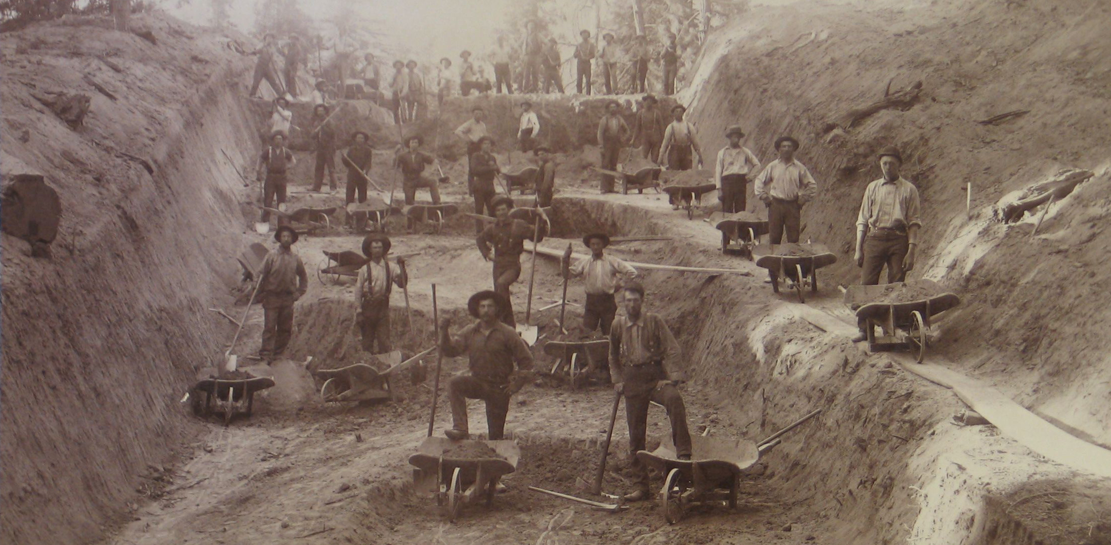 A historic black and white photo showing more than two dozen men with wheelbarrows and shovels standing in a road cut through a hill in the forest