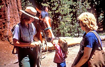 Park ranger with a horse. Girl with mother is patting the horse's nose.