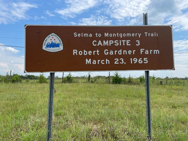 A brown highway sign reads, "Selma to Montgomery Trail. Campsite 2. Robert Gardner Farm. March 23, 1965."